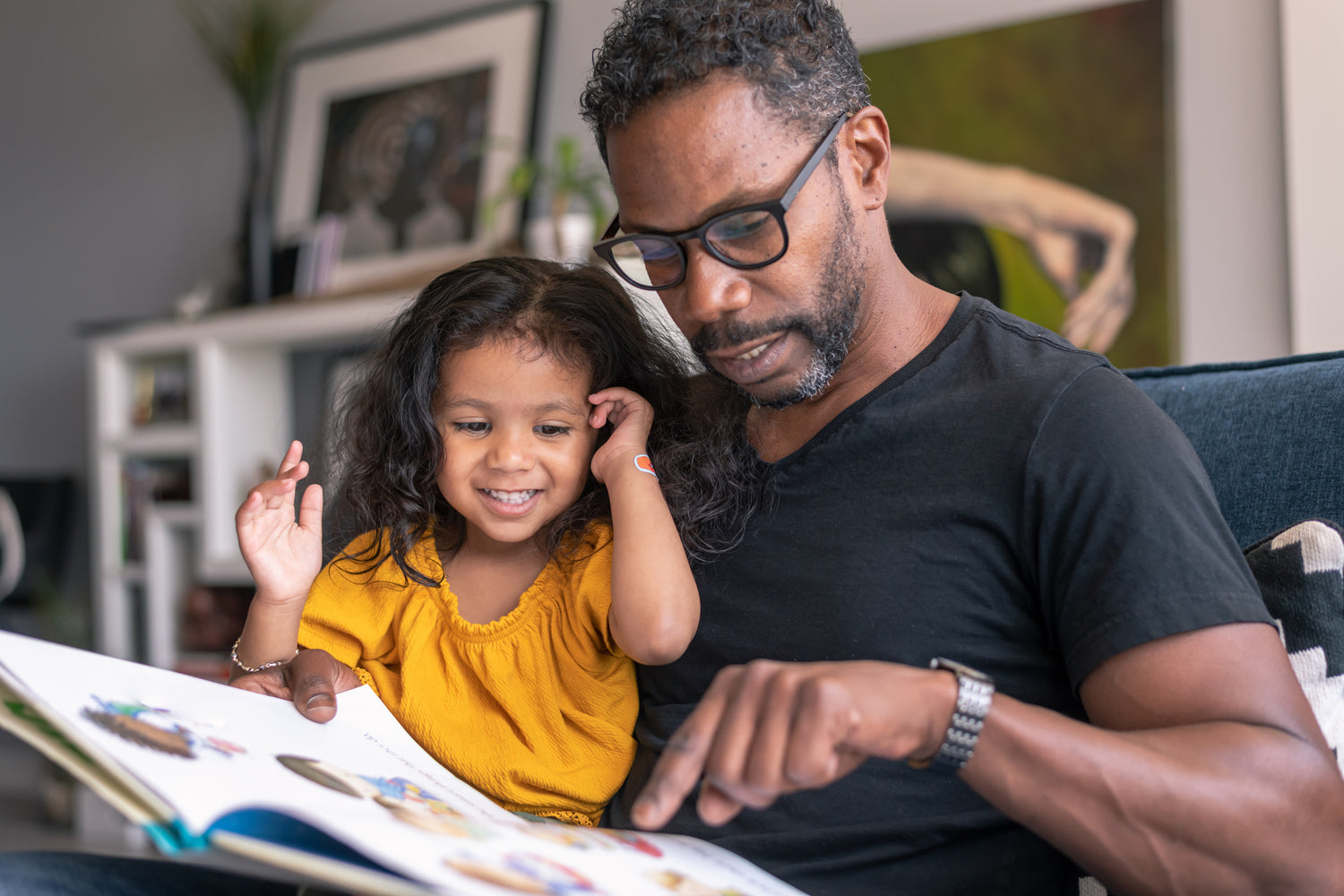 Father and toddler daughter sit on sofa smiling and reading a picture book together - creating a special bonding moment with a kids book club designed for children ages 0-8 years.
