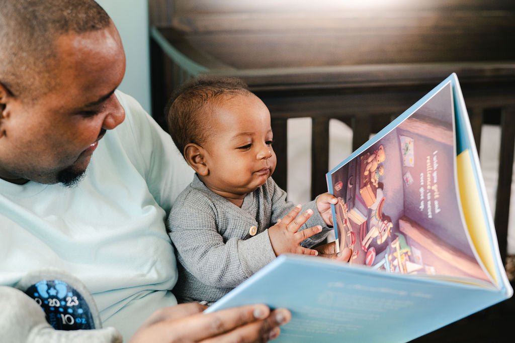 Infant son sitting on father's lap and reading a hardcover picture book. Customize your baby's book selection and build a unique library for your baby from the start.