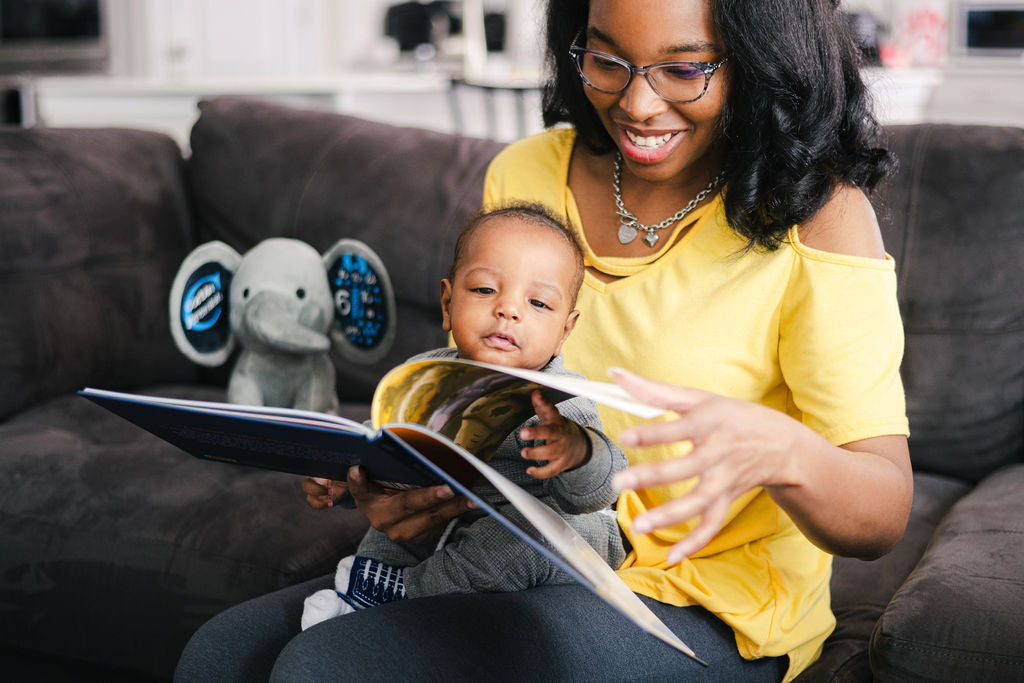Infant son sitting on mother's lap on a sofa and turning the page to a hardcover picture book. A custom, personalized birth announcement plush sits beside them on the sofa with baby's birth stats printed from a baby book club.